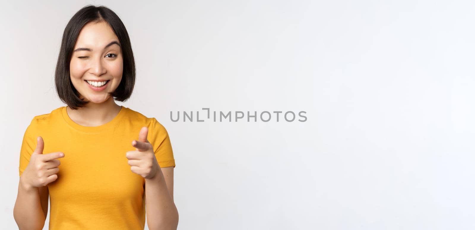Young korean girl student, pointing fingers at camera and smiling, congratulating you, choosing, inviting people, standing over white background.