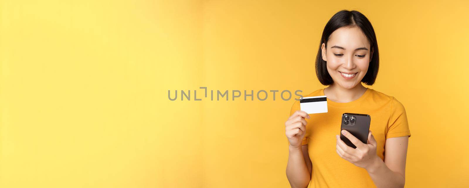 Online shopping. Smiling asian girl using credit card and mobile phone app, paying contactless, order on smartphone application, standing over yellow background by Benzoix