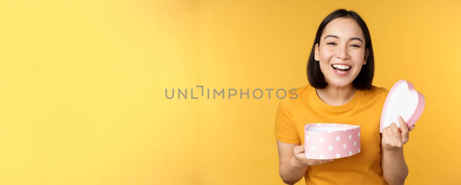 Portrait of excited asian woman, open gift box with surprised happy face, standing over yellow background.