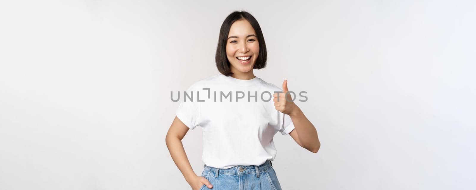 Happy beautiful korean woman, smiling pleased, showing thumbs up in approval, recommending brand or company, standing over white background by Benzoix