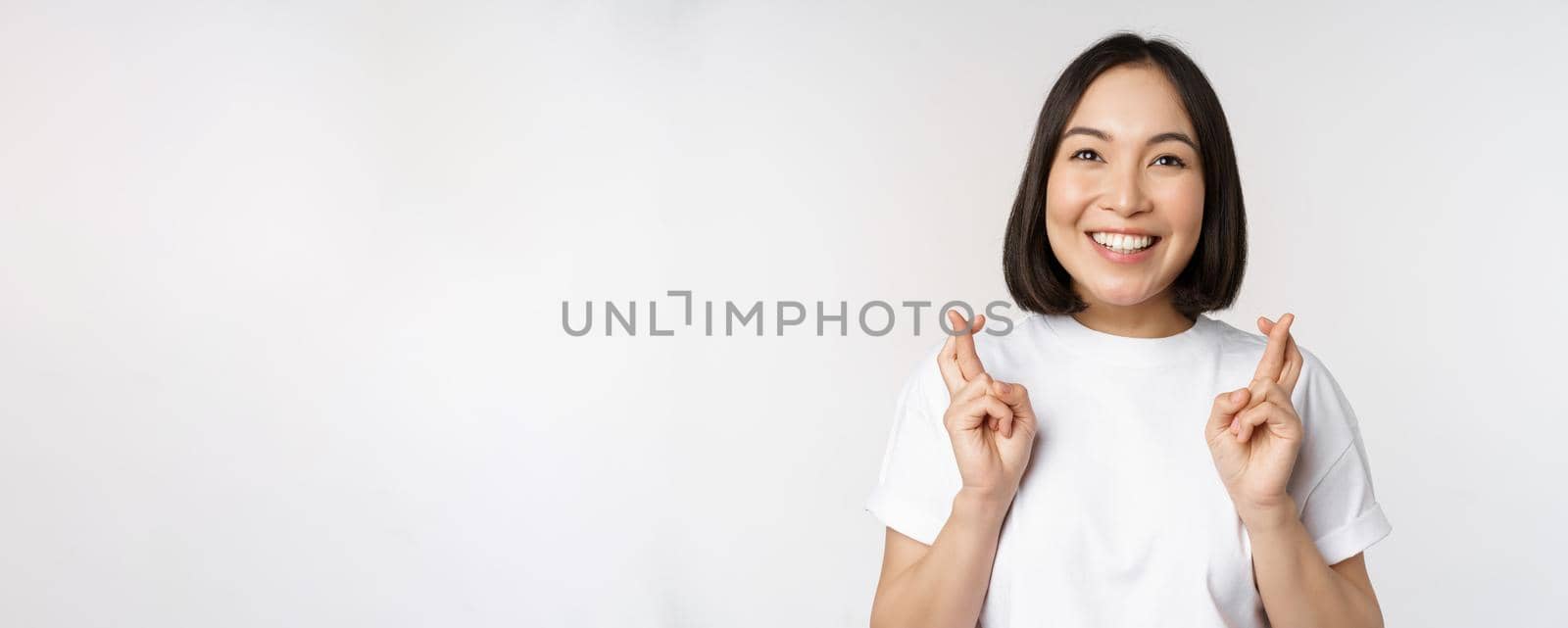 Hopeful asian girl cross fingers, making wish, anticipating, wishing smth, standing in tshirt over white background. Copy space