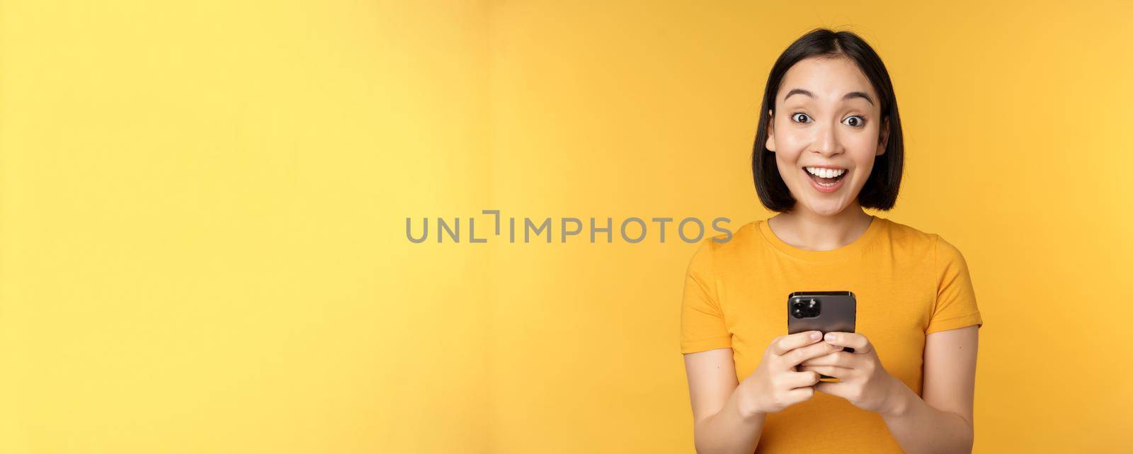 Happy asian girl smiling, standing with black mobile phone, standing against yellow background. Copy space