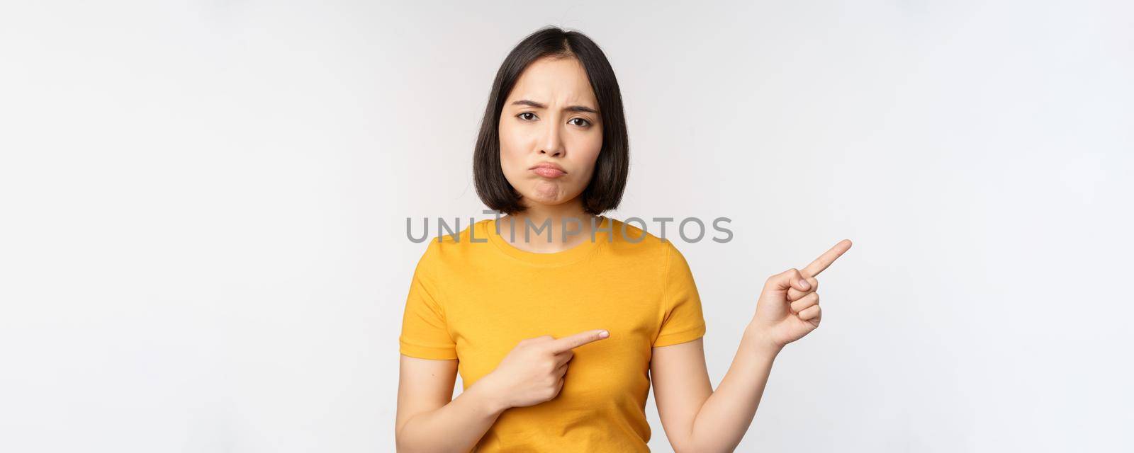 Portrait of disappointed, moody asian girl, pointing fingers right and looking at smth unfair, complaining, standing over white background.