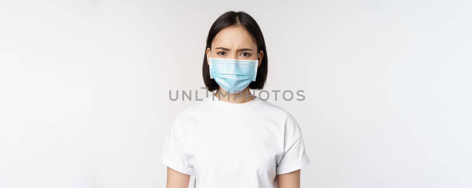 Health and covid pandemic concept. Angry young asian girl in face medical mask, frowning displeased, standing over white background.