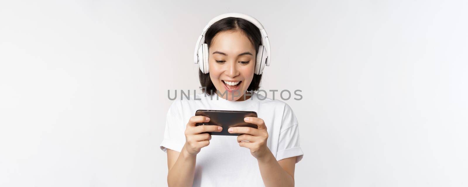 Happy asian woman in headphones, looking at smartphone, watching video on mobile phone and smiling, standing over white background.