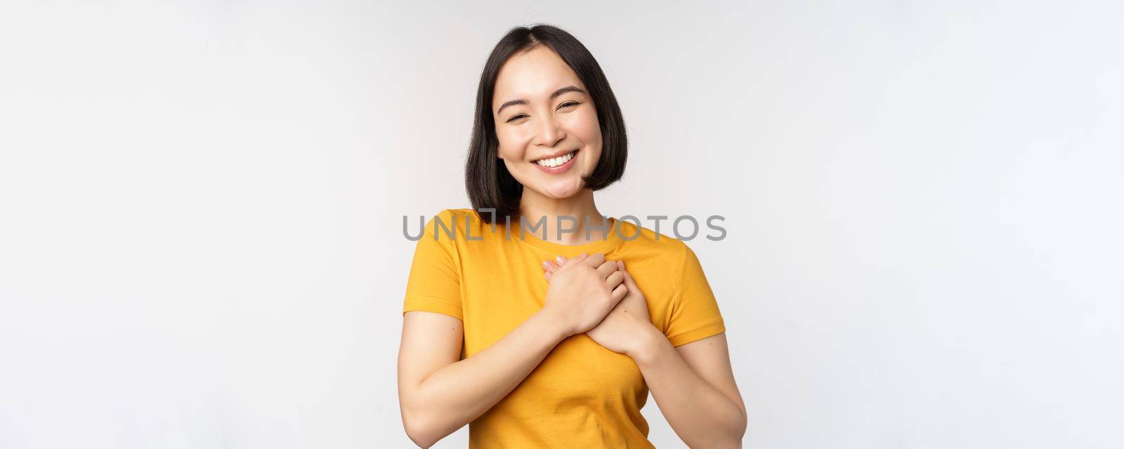 Romantic asian girfriend, holding hands on heart, smiling with care and tenderness, standing in yellow tshirt over white background by Benzoix