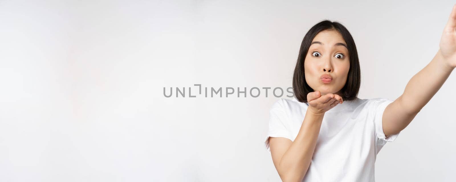 Beautiful young asian woman smiling, looking at camera, holding device, taking selfie, video chat, standing in tshirt over white background.