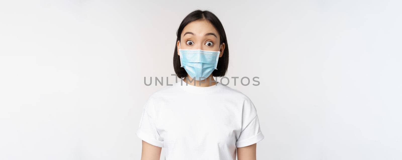 Health and covid pandemic concept. Image of korean woman in face medical mask looking surprised, reacting amazed, standing over white background.