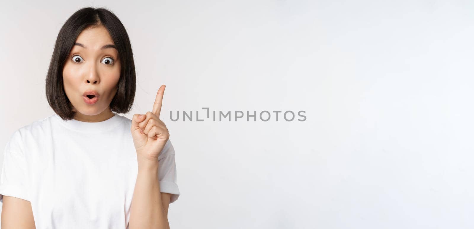 Close up of creative asian girl raising finger, suggesting smth, has an idea, pointing up, eureka sign, standing over white background by Benzoix