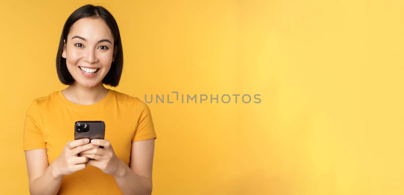 Technology. Smiling asian woman using mobile phone, holding smartphone in hands, standing in t-shirt against yellow background.