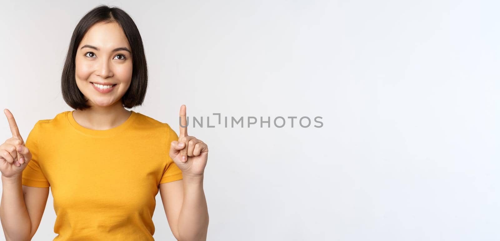 Happy beautiful asian girl showing advertisement, pointing fingers up, standing over white background.