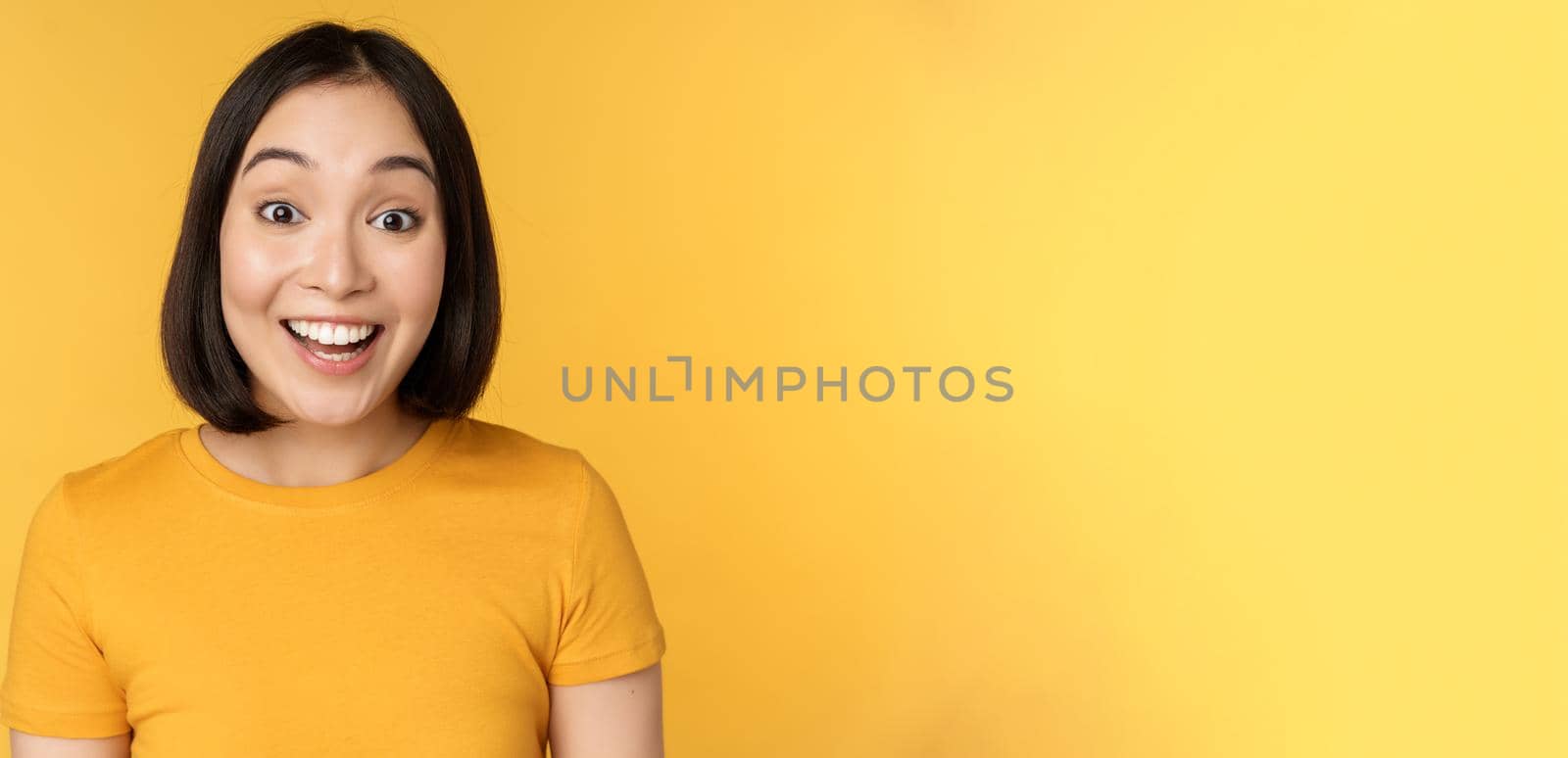 Close up portrait of asian girl showing surprised reaction, raising eyebrows amazed, reacting to big news, standing over yellow background by Benzoix