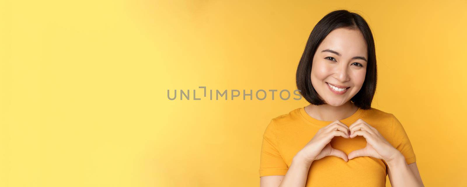 Close up portrait of smiling korean woman, showing romantic heart sign and looking happy, standing over yellow background by Benzoix