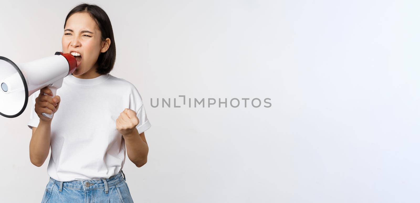 Confident asian woman shouting in megaphone, screaming and protesting. Girl activist using speaker to speak louder, standing over white background by Benzoix