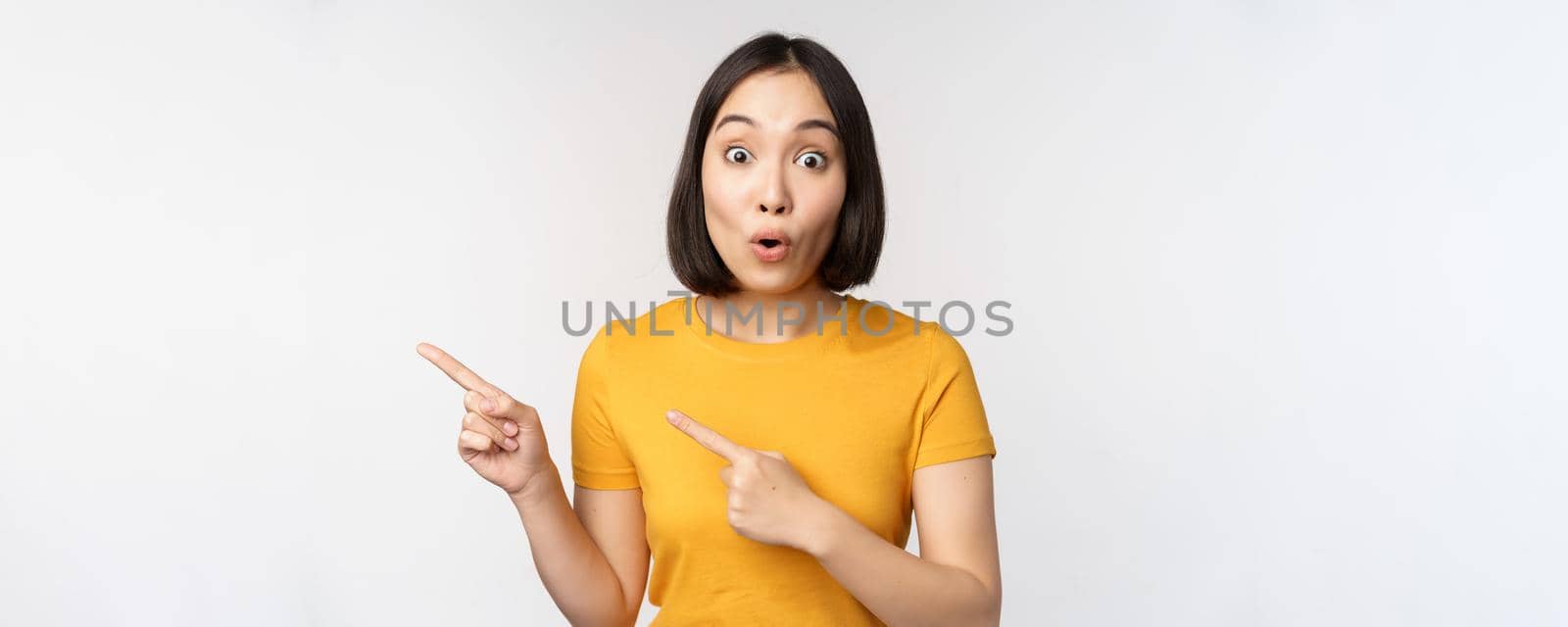 Portrait of smiling asian brunette girl in yellow tshirt, pointing fingers left, showing copy space, promo deal, demonstrating banner, standing over white background by Benzoix