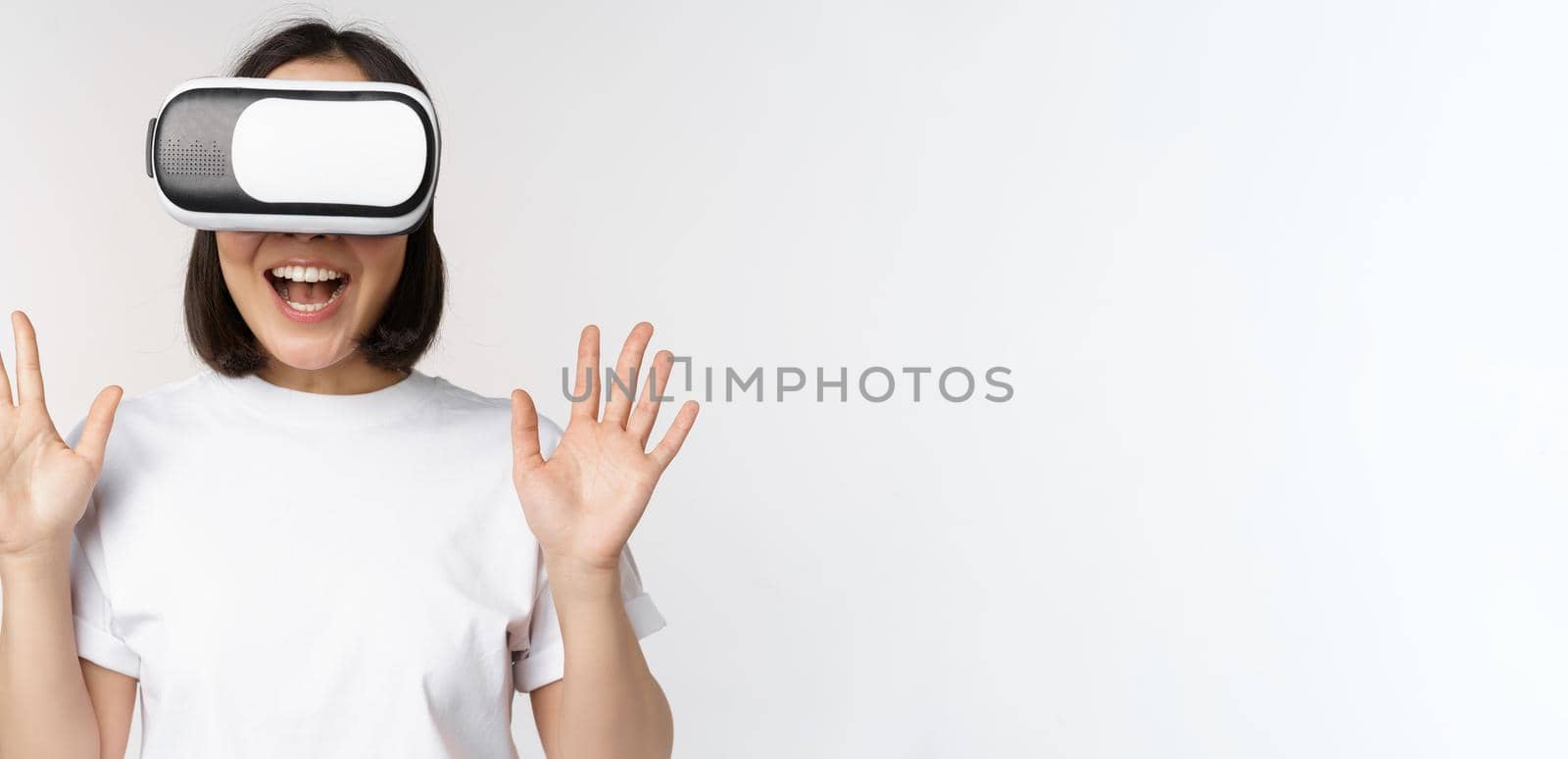 Happy asian woman using VR headset, waving raised hands and laughing, using virtual reality glasses, standing over white background.