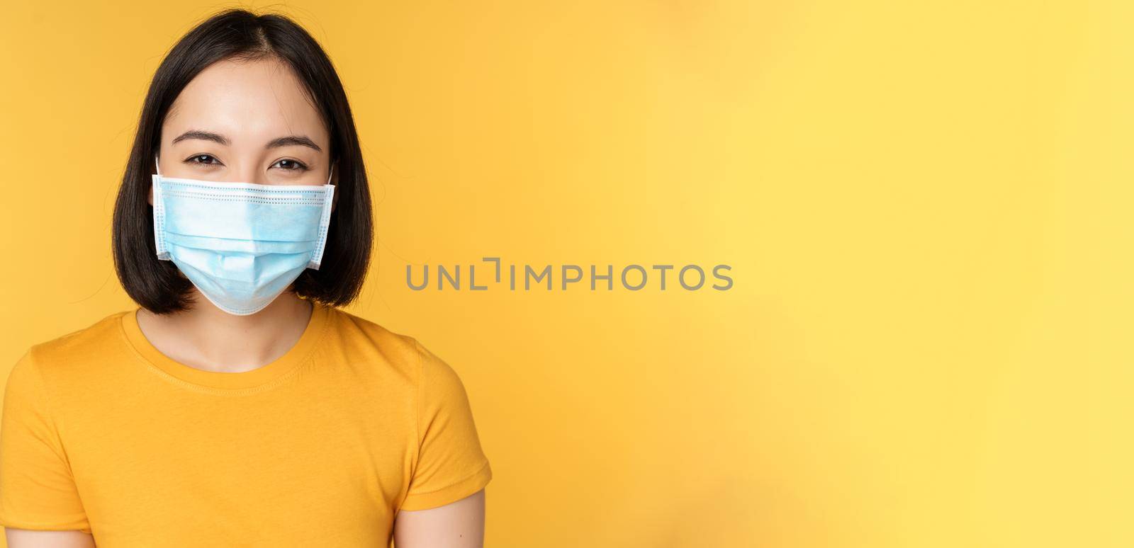 Close up of smiling happy asian woman, wearing medical face mask from covid-19, standing in yellow t-shirt over studio background by Benzoix
