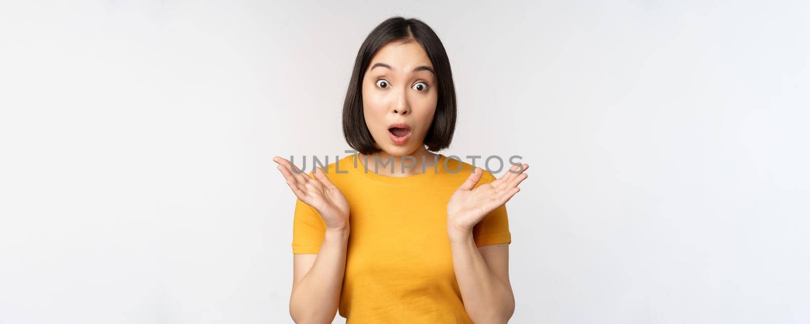 Close up portrait of asian woman looking surprised, wow face, staring impressed at camera, standing over white background in yellow t-shirt by Benzoix