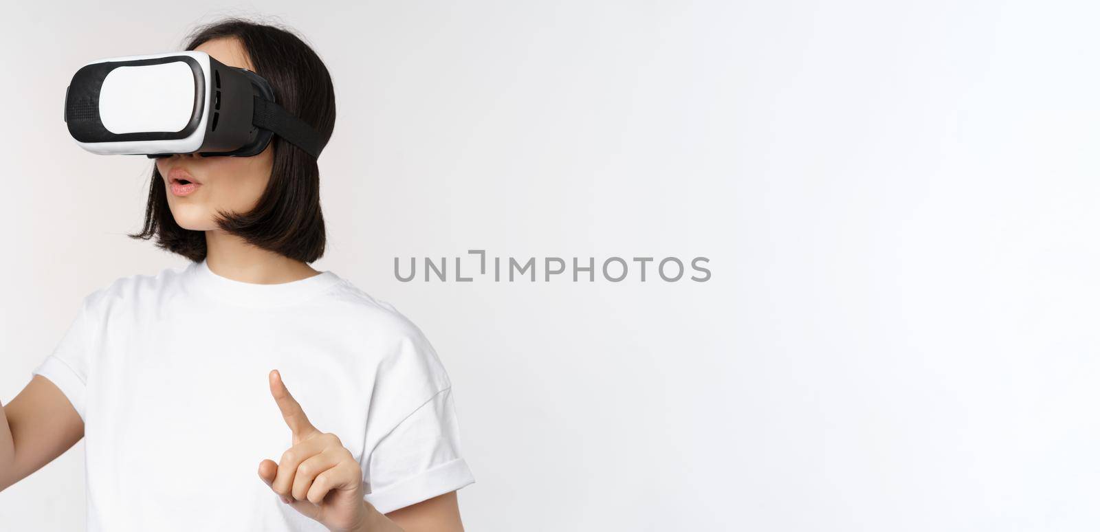 Potrait of asian woman in virtual reality glasses, pointing, choosing smth in VR headset, standing over white background by Benzoix