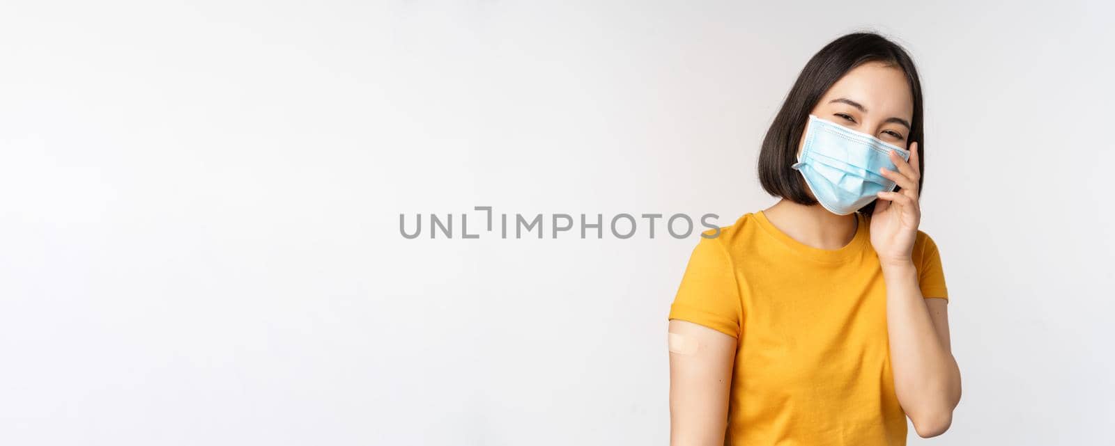 Covid-19, vaccination and healthcare concept. Cute asian girl in medical face mask, showing band aid after coronavirus vaccination, standing over white background by Benzoix