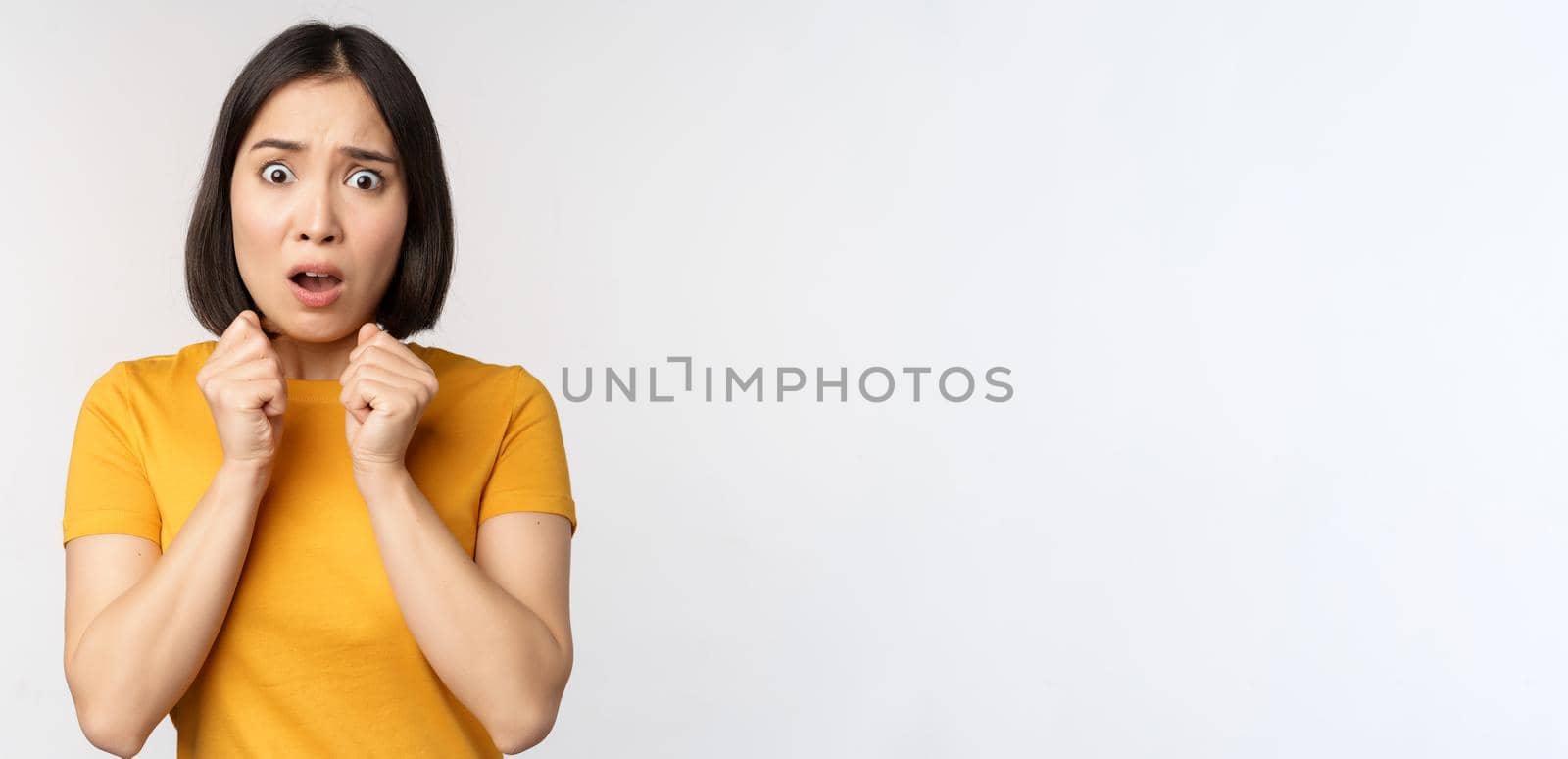 Portrait of scared asian woman shaking from fear, looking terrified and concerned, standing anxious against white background by Benzoix