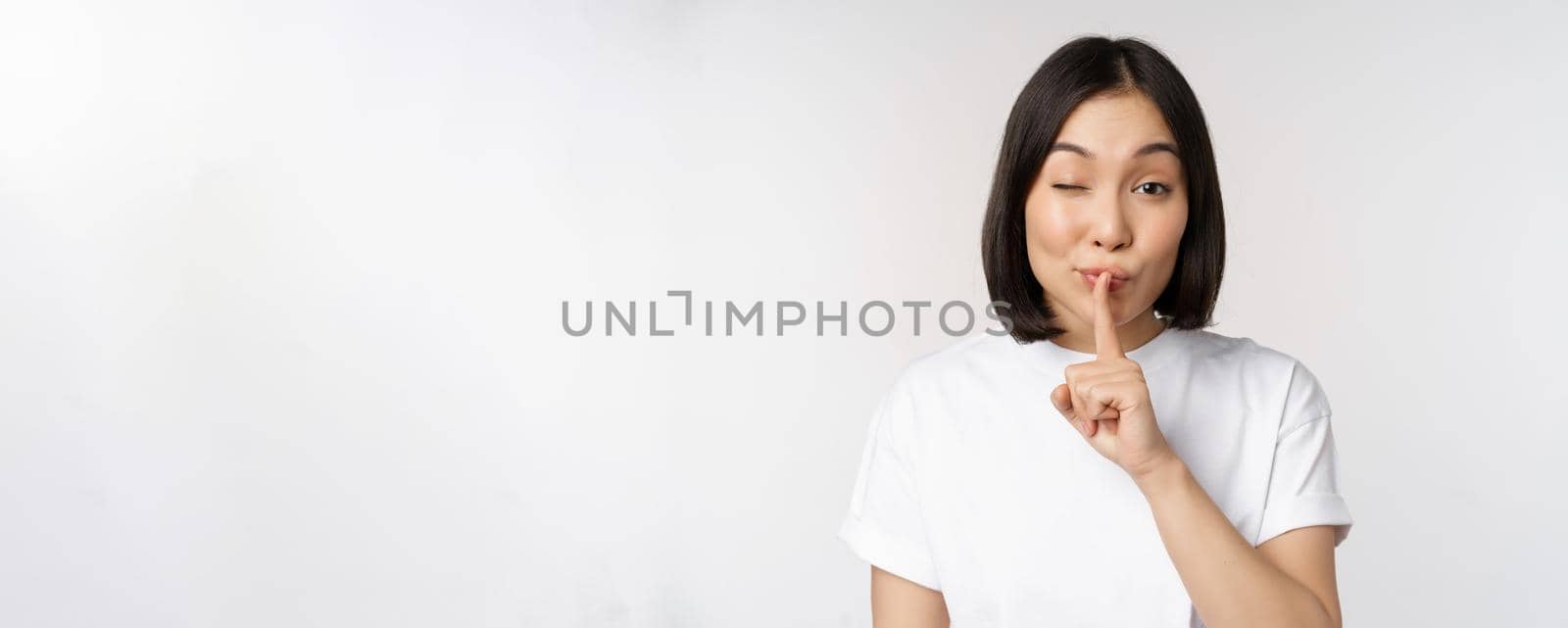 Close up portrait of young beautiful asian girl shushing, has secret, keep quiet silence gesture, press finger to lips, standing in tshirt over white background by Benzoix