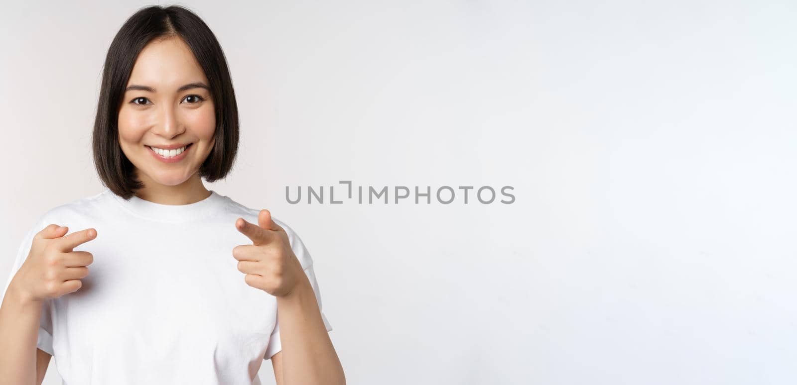 Image of smiling asian girl pointing fingers at camera, choosing, inviting you, congratulating, standing in tshirt over white background by Benzoix