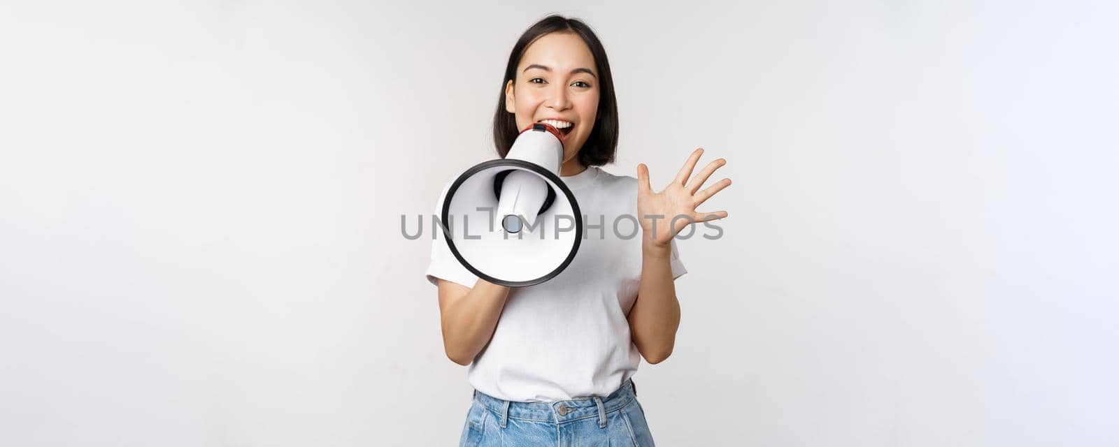 Happy asian woman shouting at megaphone, making announcement, advertising something, standing over white background.