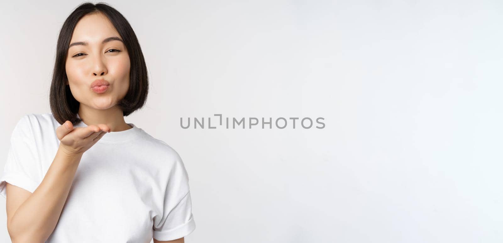 Lovely young asian woman, sending air kiss and looking coquettish at camera, standing in tshirt over white background by Benzoix