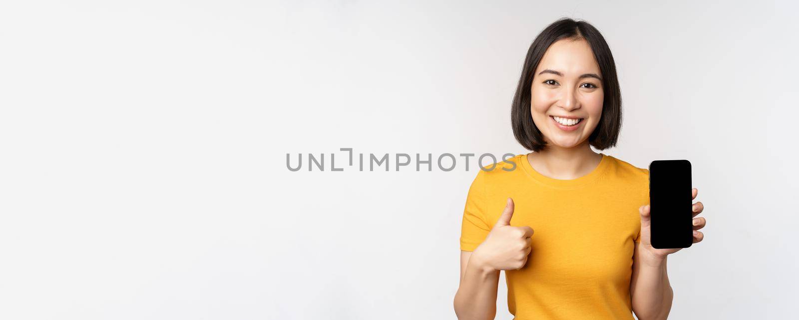 Technology and people concept. Smiling young woman showing thumb up and smartphone screen, mobile phone app interface, standing against white background.