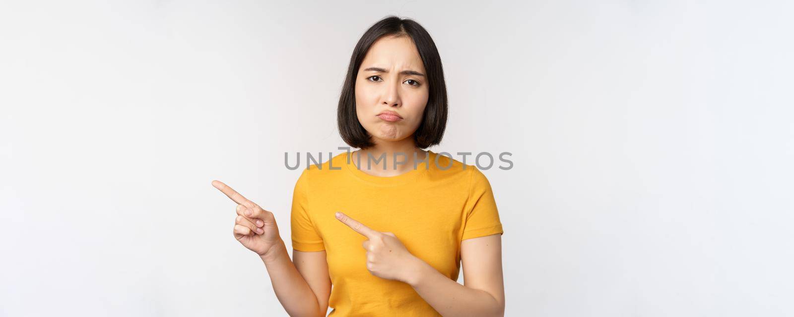 Portrait of disappointed brunette asian girl, pointing fingers left, grimacing upset, showing smth unpleasant, standing over white background by Benzoix