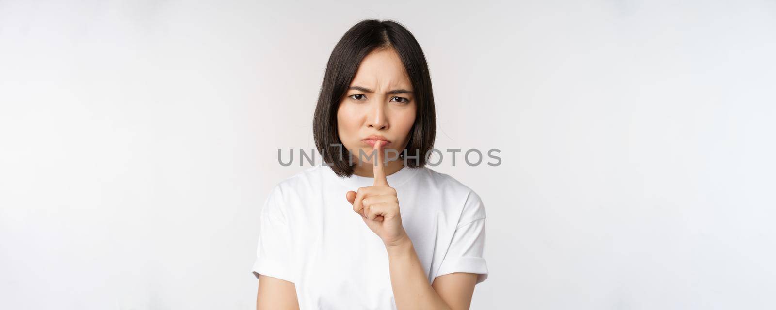 Angry asian girl shushing, keep quiet, taboo silence gesture, press finger to lips and frowning, scolding for being too loud, standing over white background by Benzoix