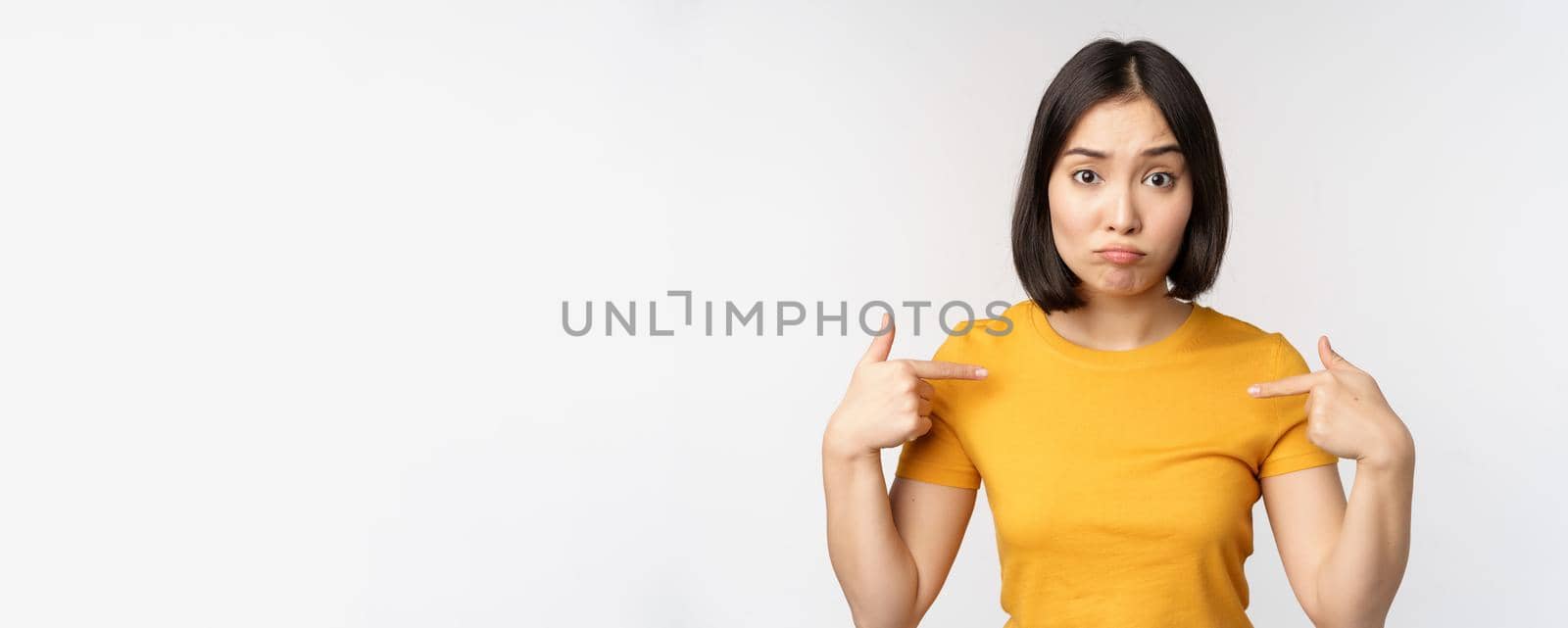 Confused asian woman pointing at herself, looking in disbelief, standing over white background. Copy space