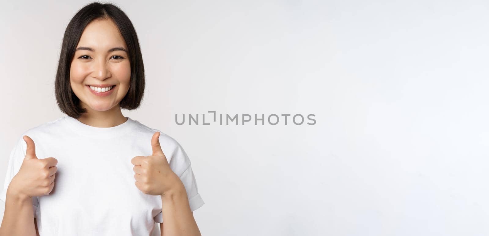 Happy young korean woman showing thumbs up and smiling, give positive feedback, recommending smth, standing over white background by Benzoix