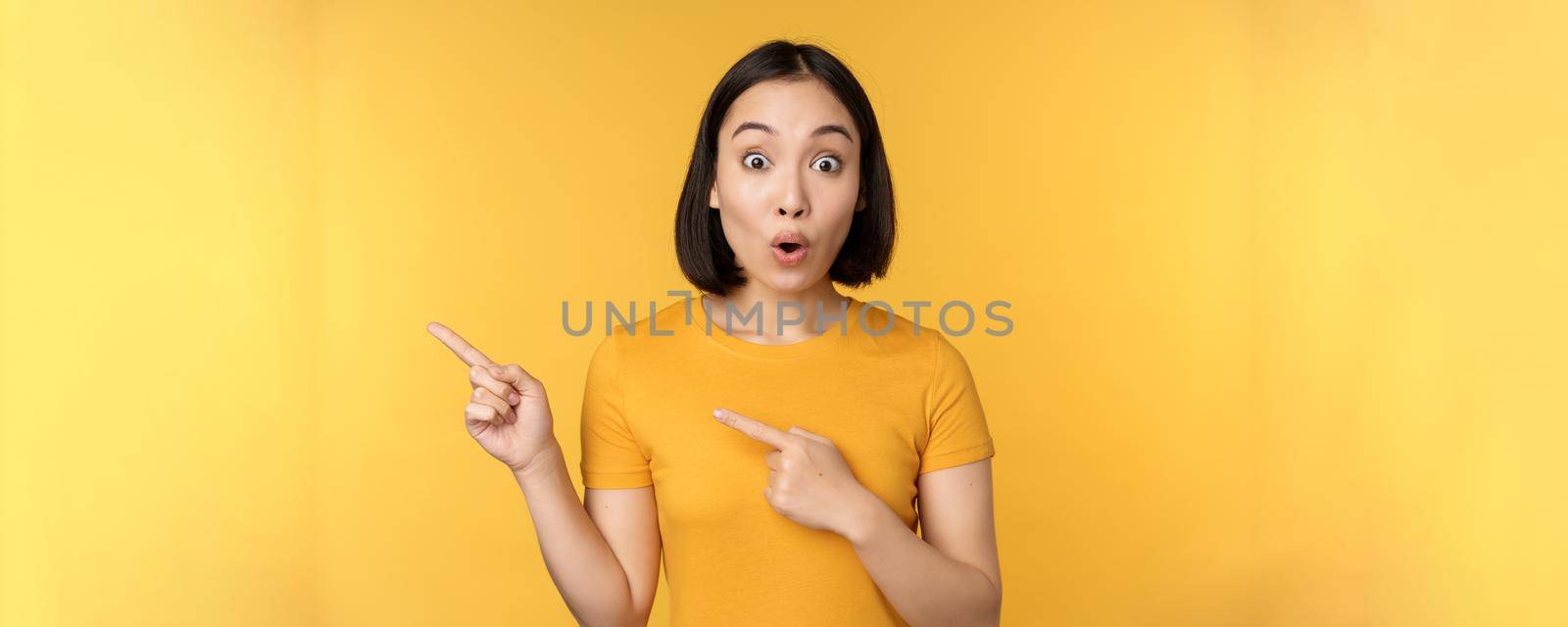 Smiling asian woman pointing fingers left, showing advertisement on empty copy space, standing over yellow background.