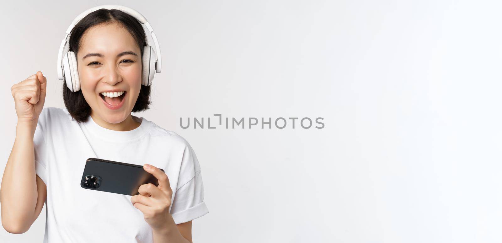 Happy asian girl gamer, playing on mobile phone, watching on smartphone, wearing headphones, standing over white background.