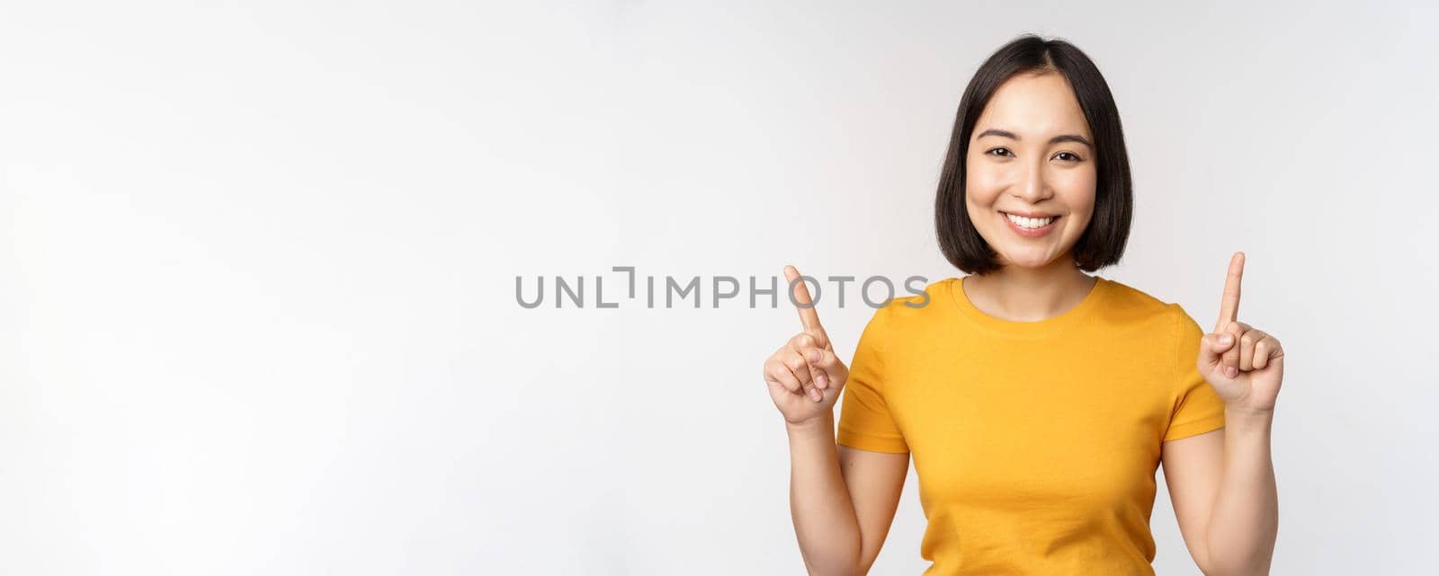 Portrait of beautiful japanese girl smiling, pointing fingers up, showing advertisement, standing in yellow tshirt against white background.