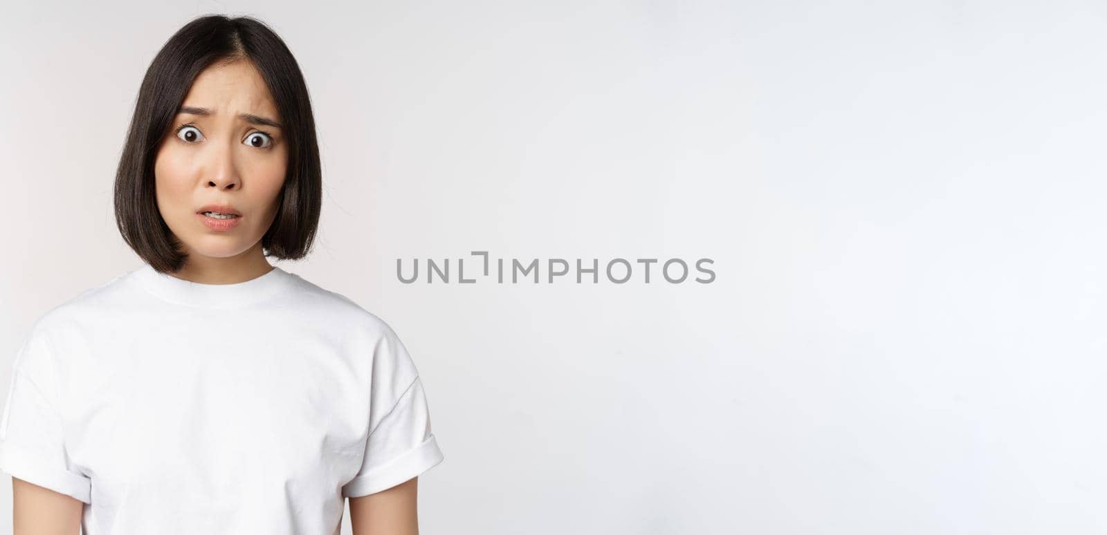 Image of shocked and frustrated asian woman, looking alarmed at camera, cant understand, puzzled, standing over white background by Benzoix