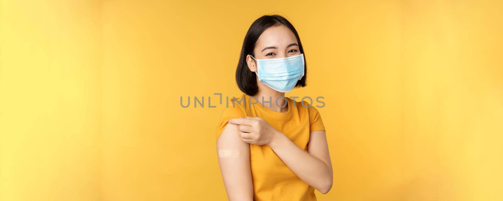 Vaccination and covid-19 pandemic concept. Smiling asian woman in medical face mask, showing her shoulder with band aid after vaccinating from coronavirus, yellow background by Benzoix