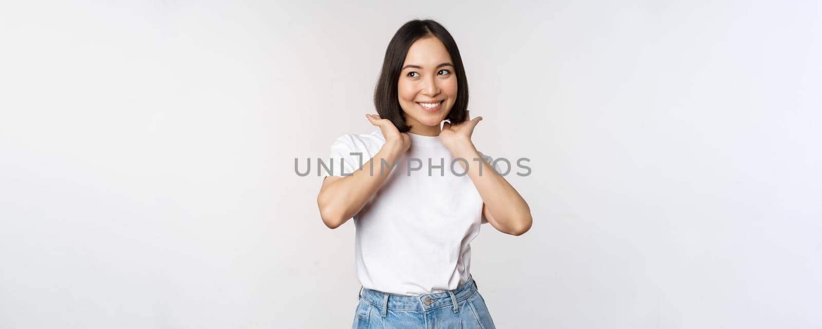 Portrait of cute, beautiful asian woman touching her new short haircut, showing hairstyle, smiling happy at camera, standing over white background.