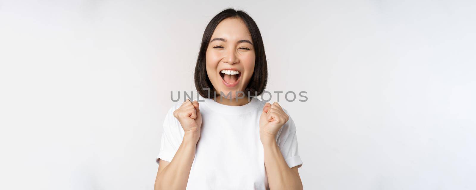Portrait of enthusiastic asian woman winning, celebrating and triumphing, raising hands up, achieve goal or success, standing over white background by Benzoix