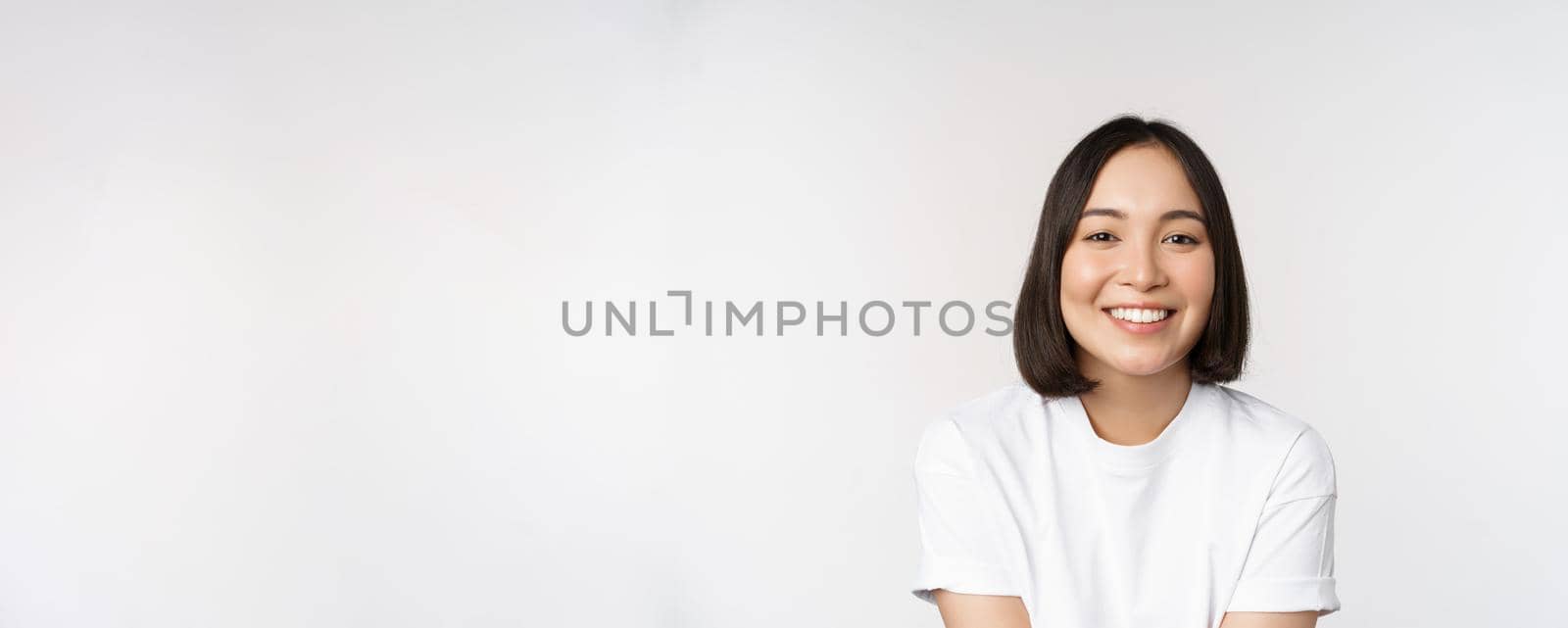 Beautiful korean girl smiling, white teeth, looking lovely at camera, standing in white tshirt over studio background.