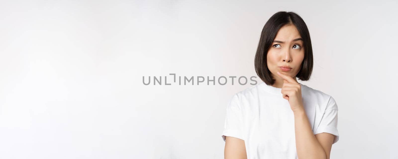 Thoughtful asian woman looking aside, pondering, making assumption, thinking or choosing smth, standing over white background by Benzoix