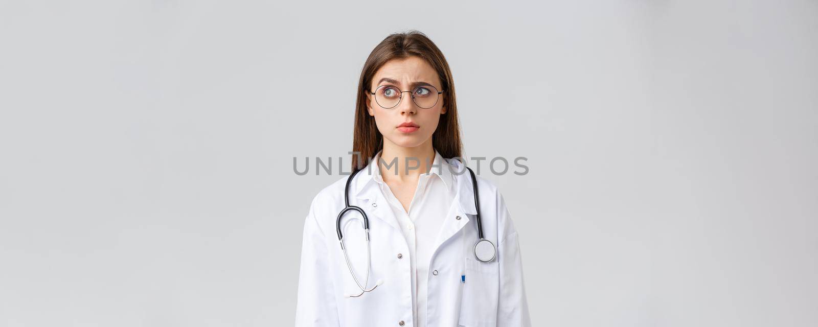 Healthcare workers, medicine, insurance and covid-19 pandemic concept. Doctor see something strange. Confused and suspicious female doctor in white scrubs, glasses, frowning and staring left by Benzoix