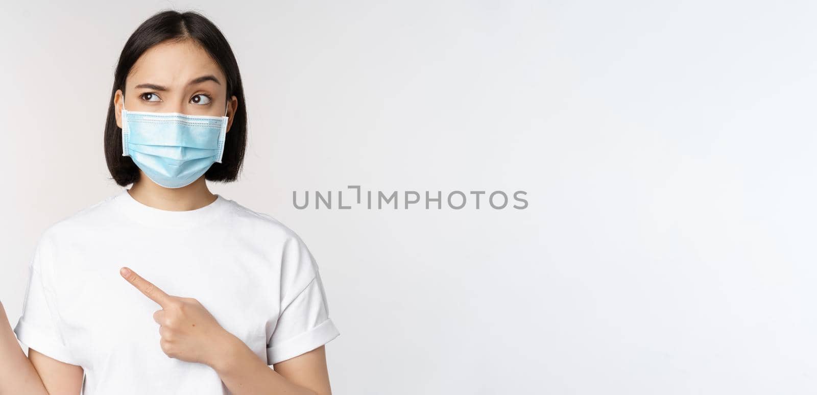 Image of asian woman in medical mask from covid, looking confused left, pointing at logo, showing advertisement, standing over white background by Benzoix
