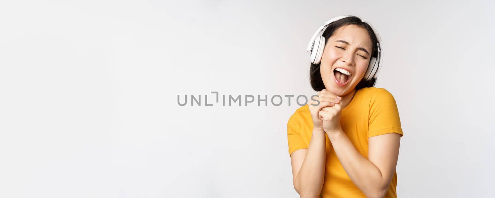 Happy asian girl dancing, listening music on headphones and smiling, standing in yellow tshirt against white background. Copy space