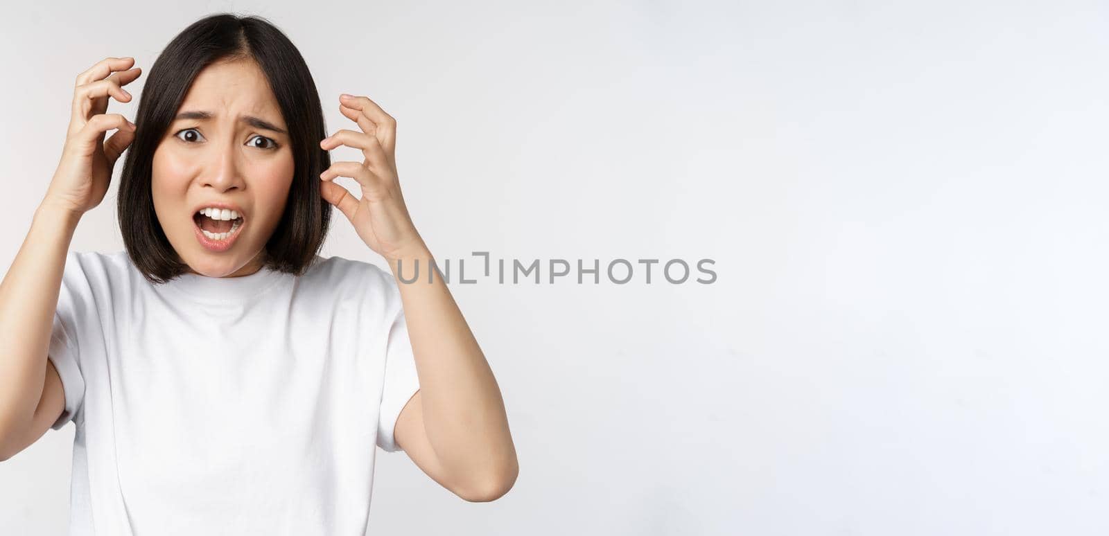 Korean woman freaking out, shaking hands and shouting in panic, screaming worried, standing over white background.