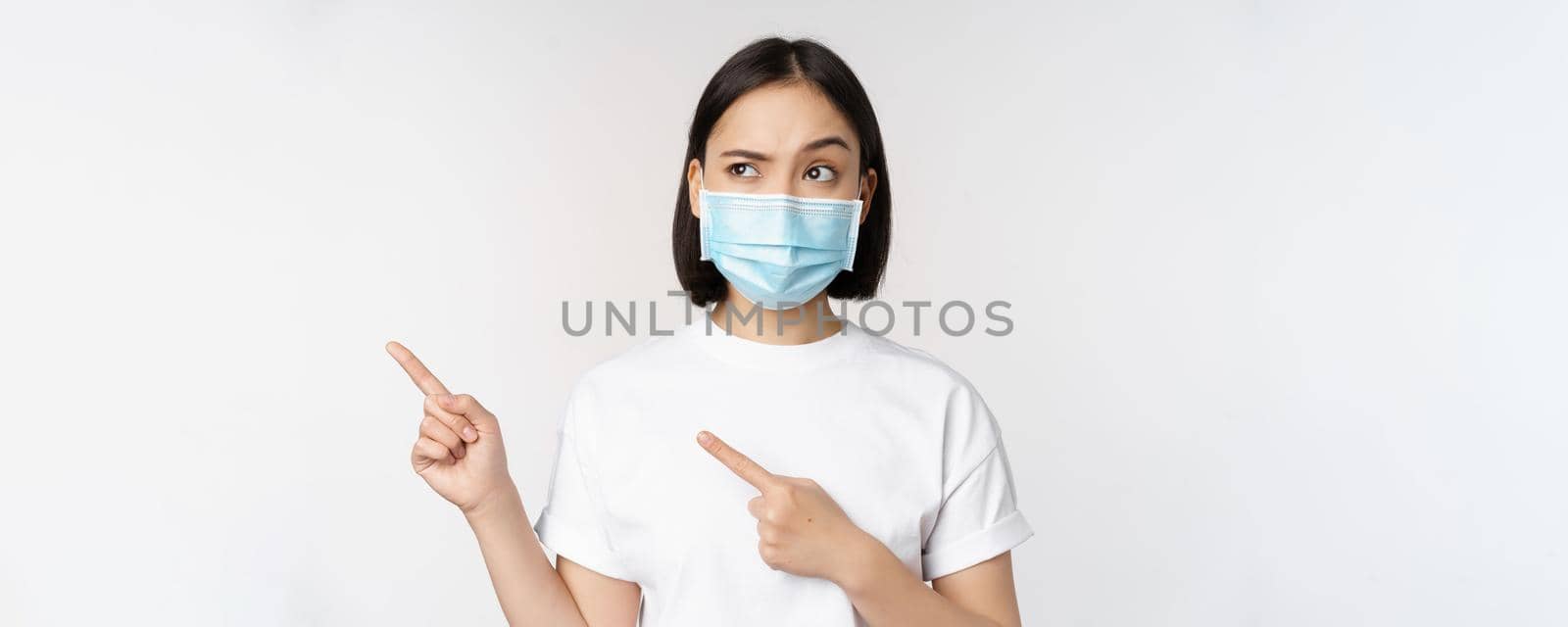 Image of asian woman in medical mask from covid, looking confused left, pointing at logo, showing advertisement, standing over white background by Benzoix