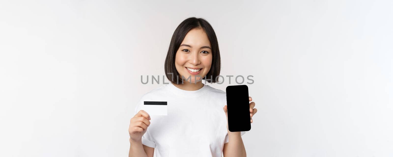 Portrait of smiling young asian woman showing mobile phone screen and credit card, standing over white background by Benzoix