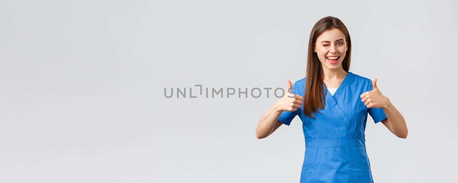 Healthcare workers, prevent virus, insurance and medicine concept. Optimistic good-looking female nurse or doctor in scrubs, wink encourage visit clinic, show thumbs-up in approval.